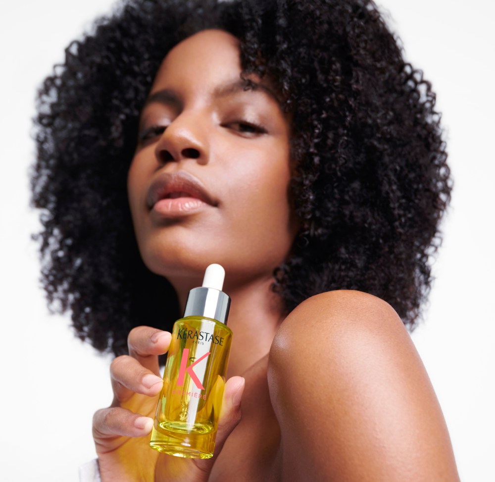 Ebonee Davis con su precioso cabello rizado sostiene el mejor aceite para cabello dañado, Première Huile Gloss Réparatrice.
