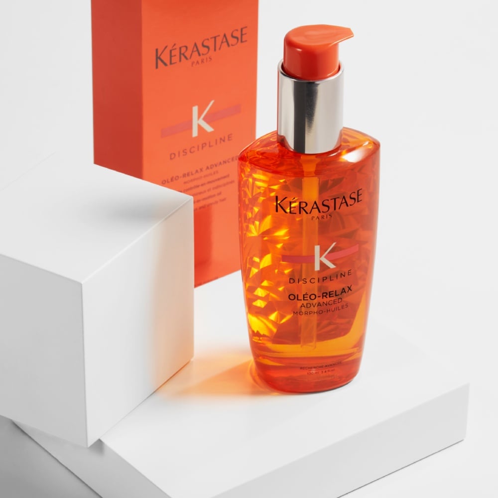 El aceite capilar Discipline Oleo Relax se encuentra sobre una sencilla plataforma blanca con un fondo gris puro, mientras que su brillante color naranja resplandece