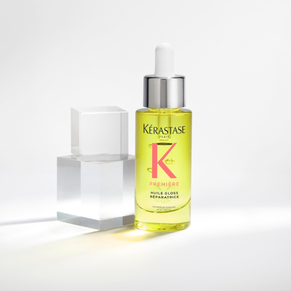 El aceite capilar Première Huile gloss réparatrice se muestra sobre un fondo gris puro con cubos transparentes. El aceite repara el brillo y las puntas abiertas a la vez que neutraliza la opacidad.