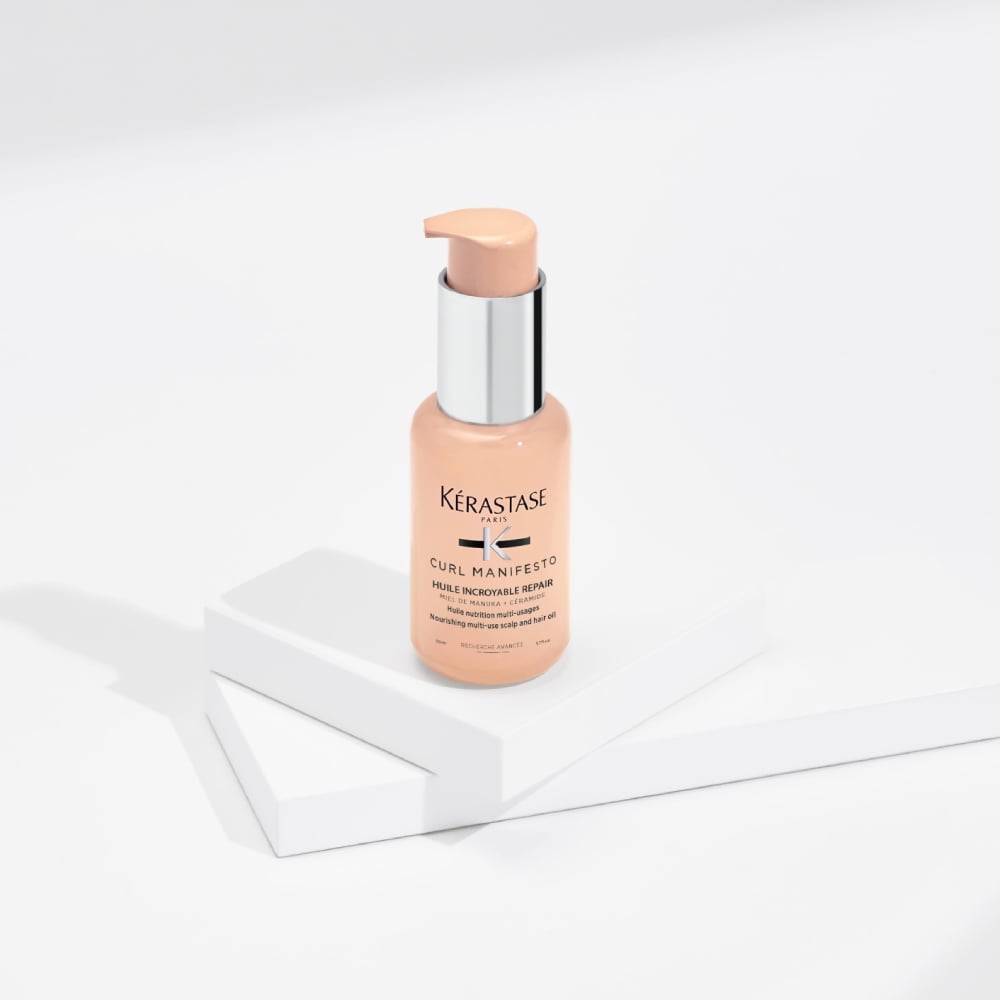 El aceite capilar Curl Manifesto Huile Sublime Repair se muestra sobre un fondo gris puro con cubos transparentes. Este aceite hidrata y define la forma del cabello rizado al tiempo que reduce el frizz y el quiebre.